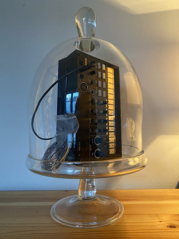 Korg drum machine sealed under a blown glass cloche on a glass stand